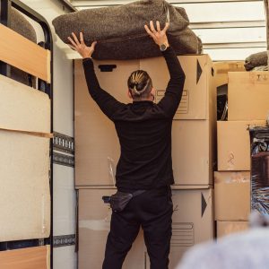 Caucasian worker with his back and ponytail placing a bundle on top of cardboard boxes inside a moving truck.