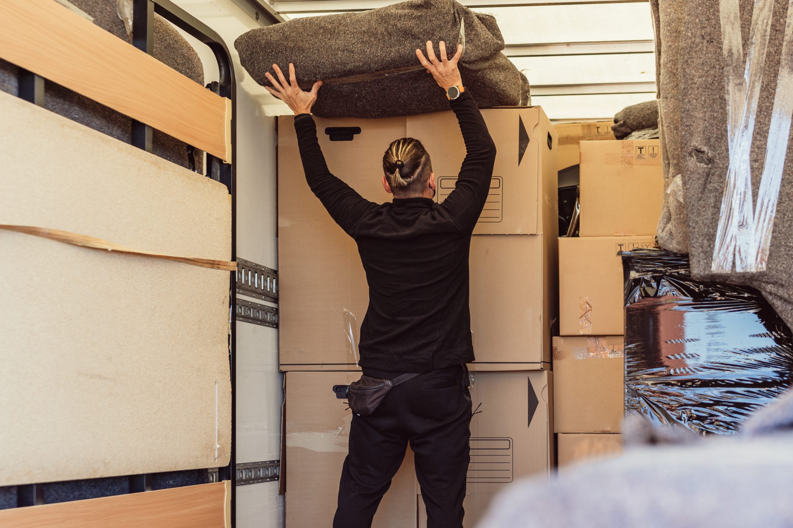 Caucasian worker with his back and ponytail placing a bundle on top of cardboard boxes inside a moving truck.